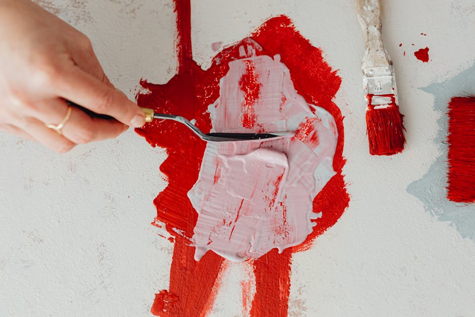 Hand using palette knife to mix red and white paint on canvas, demonstrating artistic technique.