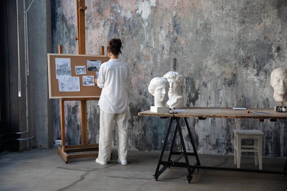 A woman studies sketches on a corkboard in a rustic art studio with sculptures and a textured wall.